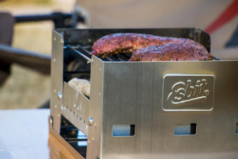 ESBIT fold-away compact BBQ charcoal grill with two steaks cooking on the grill outdoors.