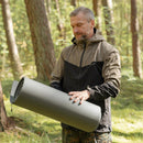 Man outdoors holding a rolled-up original British military gray sleeping pad in a forest setting