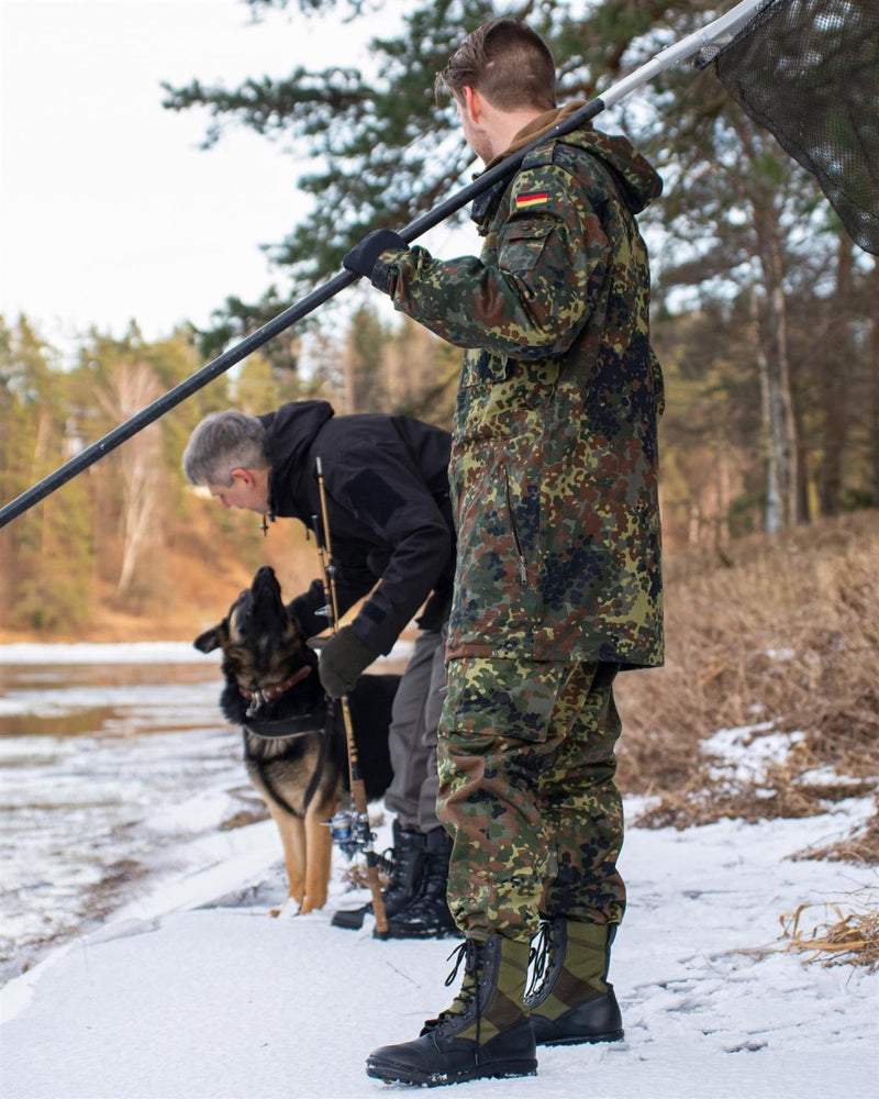 Man in German camouflage uniform wearing black and OD green Bates side zip military boots in snowy outdoor setting.
